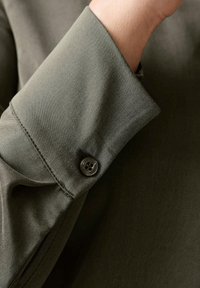 Dunkelolivgrünes Langarmhemd mit einem Umschlag, der einen einzelnen schwarzen Knopf und eine glatte Stoffstruktur aufweist. Dezente Nahtdetails sind sichtbar.