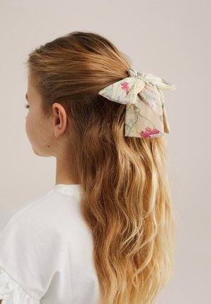 Fată cu păr blond lung și ondulat prins în spate cu o fundă din material crem, cu imprimeu floral colorat, purtând un top alb.