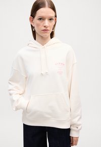 Felpa color crema realizzata in un materiale morbido, con una tasca frontale, lacci e un dettaglio del logo rosa sul petto. Jeans scuri sotto.