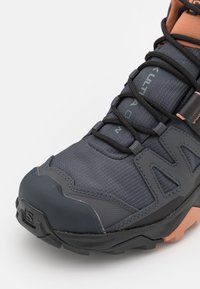 Scarpe da trekking grigie scure con una tomaia in tessuto testurizzato, punta in gomma e lacci neri. Presentano un accento arancione vicino alla caviglia e una suola con motivo.