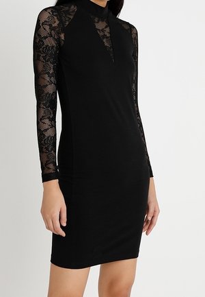 Femme portant une robe noire ajustée avec des manches longues en dentelle et un détail en dentelle en forme de V sur la poitrine.
