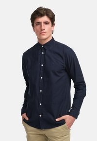 Chemise bleu marine à boutonnières avec un col évasé, boutons blancs et un petit emblème sur la poitrine. Le tissu a une texture lisse.