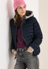 Veste matelassée bleu marine avec une capuche grise, portée sur un pull bordeaux, associée à un jean gris. Le modèle porte un bonnet en maille rose pâle.