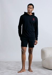 Μαύρο φούτερ με κουκούλα και μακριά μανίκια με κόκκινο λογότυπο καρδιάς, συνδυασμένο με μαύρο σορτς. Το ύφασμα φαίνεται μαλακό· και τα δύο ρούχα έχουν άνετη εφαρμογή.