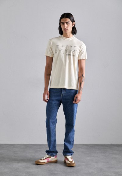Beige cotton T-shirt with graphic print and text, paired with blue denim jeans. Brown and pink sneakers complete the outfit.