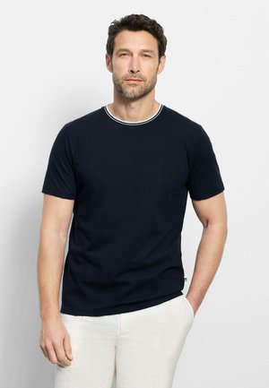 Uomo con capelli castani corti e barba indossa una maglietta nera a maniche corte e pantaloni bianchi, in piedi davanti a uno sfondo chiaro e uniforme.