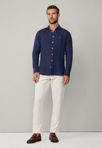 Navy long-sleeve linen shirt with a button-down collar, featuring a subtle front logo. Paired with white trousers and brown loafers.