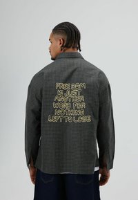 Grijze geruite blouse met lange mouwen, met gele tekst op de achterkant: "VRIJHEID IS GEWOON EEN ANDER WOORD VOOR NIKS MEER TE VERLIEZEN."