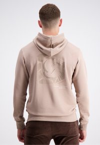 Bež kapuca s teksturiranim grafičkim dizajnom na leđima. Ima kabel za zatezanje na kapuljači i rebraste manžete. Mekani pamučni materijal.
