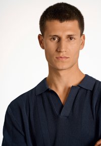 Merino-poolo paita tummansinisellä värillä, jossa on pystysuuntainen joustin, lyhyet hihat ja V-kaulus. Sileä pinta ja istuva leikkaus.