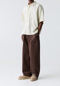 Jasnożółta, krótko rękawowa koszula z guzikami w pionowe paski, zestawiona z luźnymi, brązowymi spodniami z bocznymi kieszeniami i beżowymi butami.