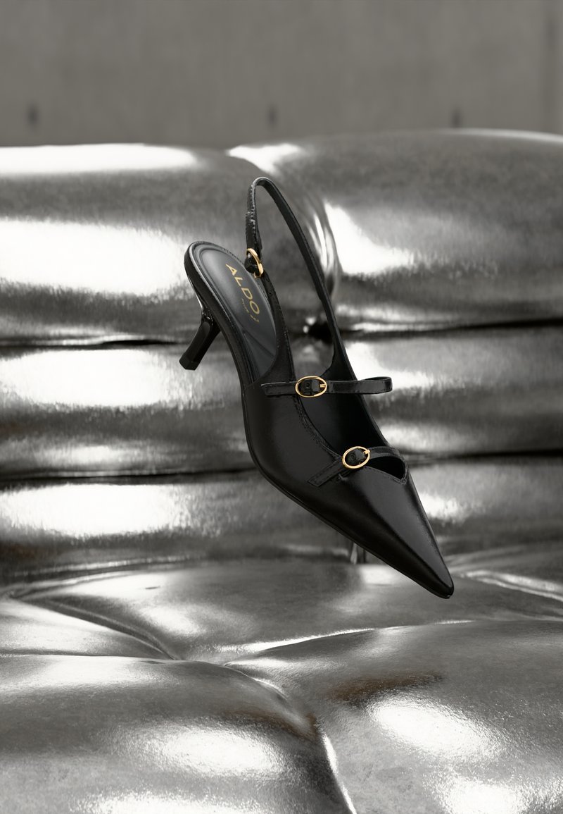 Escarpin slingback noir à bout pointu avec deux brides à boucle dorée reposant sur une surface rembourrée argentée brillante.