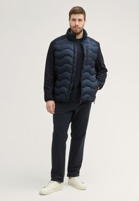 Gilet rembourré marine avec un col haut et un motif de matelassage en zigzag, superposé sur un pull sombre et un pantalon foncé, complété par des baskets blanches.