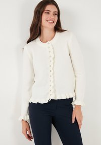 Weißer, gestrickter Cardigan mit gewelltem Knopfverschluss, rundem Ausschnitt und Rüschenabschluss an Saum und Ärmeln. Weicher Stoff, figurnaher Schnitt.
