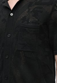 Chemise en maille noire à manches courtes avec une poche avant, une fermeture par boutons et un motif texturé complexe sur le tissu.