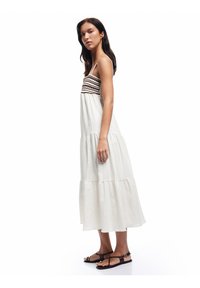 Weißes maximales Kleid mit tiered Schnitt und braunem Zickzackmuster am Oberteil sowie dünnen Trägern, kombiniert mit schwarzen Riemchensandalen.