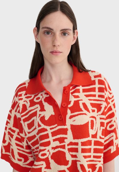 Jeune femme aux longs cheveux bruns portant un pull court à manches courtes rouge et beige à motifs avec un col boutonné sur un fond uni.