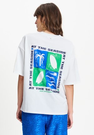 Vrouw draagt een witte oversized T-shirt met blauw-groene kustgrafiek en de tekst "Aan zee" op de achterkant en blauwe gestructureerde broek.