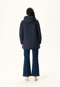 Marineblauwe pufferjas met capuchon, cinched taille en lange mouwen. Combinatie met blauwe flared jeans en beige hakken.