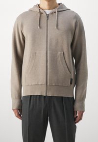 Beige Zip-Hoodie aus weichem Material mit einer Fronttasche und einer Kordelzugkapuze. Kombiniert mit anthrazitgrauen Hosen.