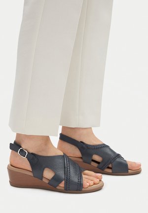 Piedi con sandali con zeppa in pelle nera e cinturini alla caviglia, abbinati a pantaloni bianchi a gamba dritta, su sfondo bianco.