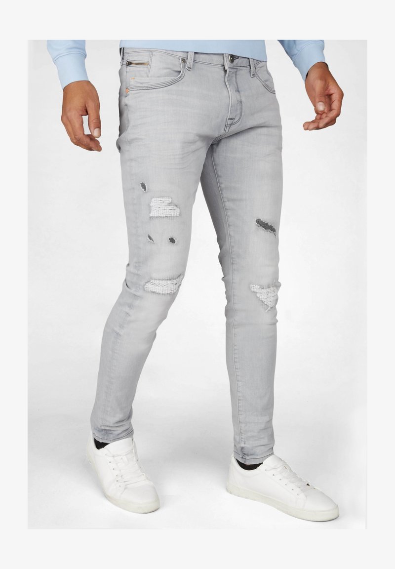 Jean skinny gris clair avec détails usés, y compris des patchs et des trous. Assorti à des baskets blanches, affichant un look décontracté.
