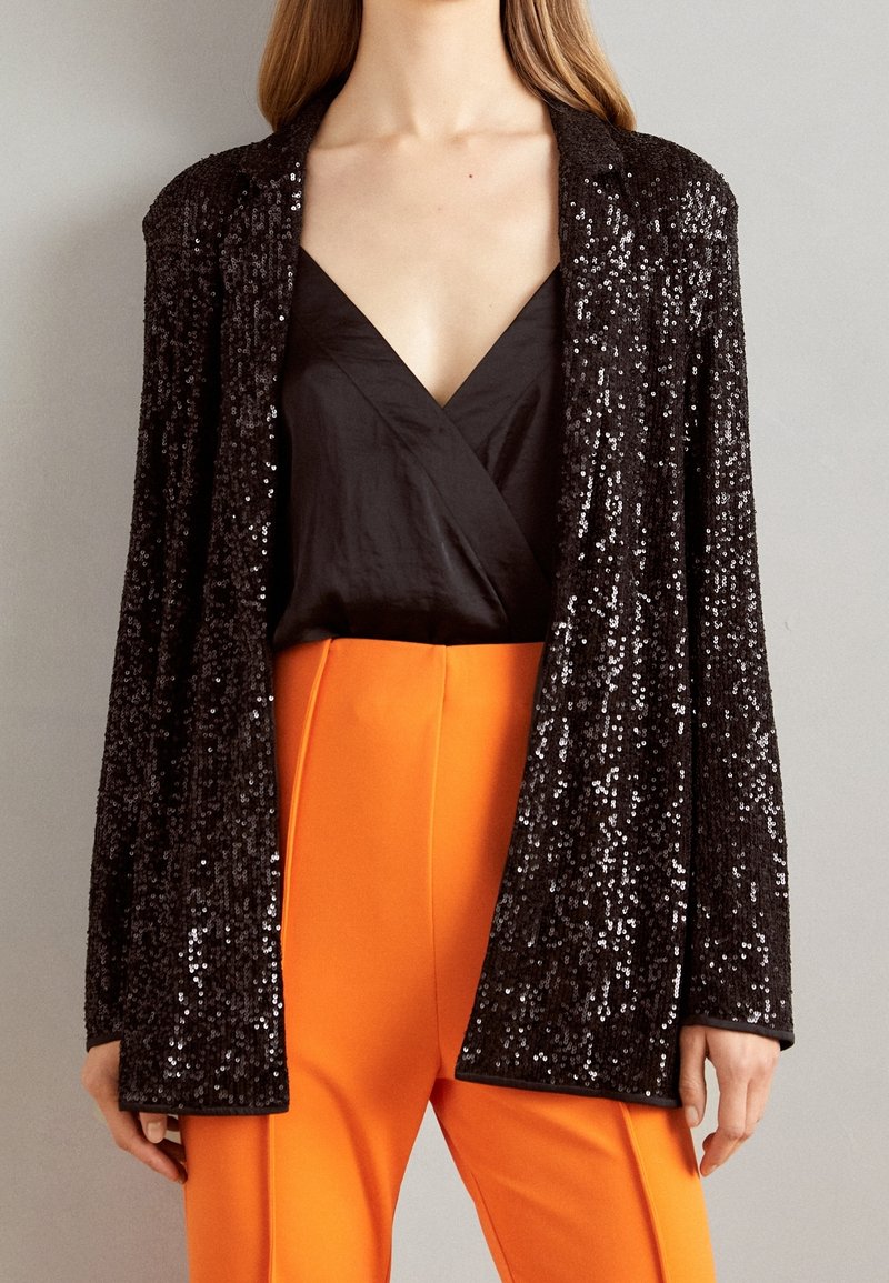 Blazer negro con lentejuelas y acabado texturizado, con apertura frontal y mangas largas, combinado con un top negro de escote en V y pantalones naranja brillante.