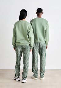 Duas pessoas vestindo sweatshirts verdes combinando e calças de treino verdes estampadas, de pé lado a lado, voltadas para fora em um chão neutro.