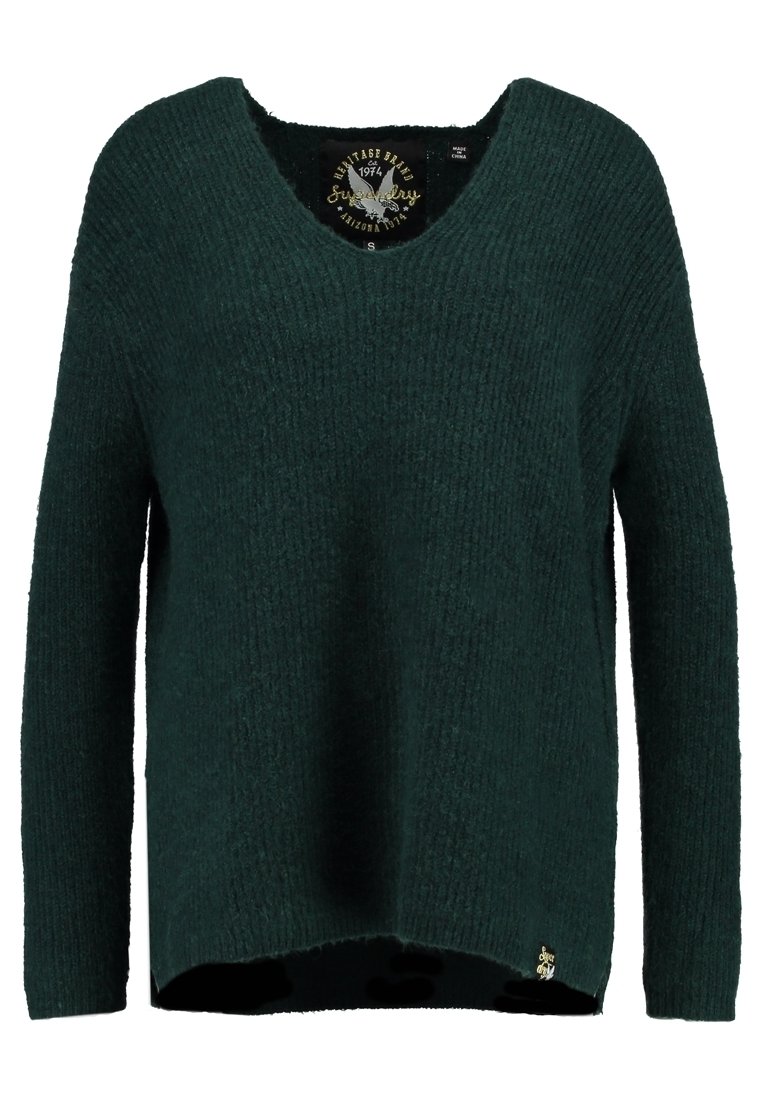 Pull en maille vert foncé à manches longues avec un col rond et une petite étiquette logo dorée près de l'ourlet inférieur.