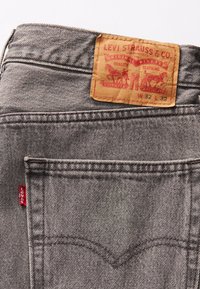 Jeans de denim gris con un parche de cuero en la cintura que presenta estampados en rojo y negro; incluyen dos bolsillos traseros y una pequeña etiqueta de logo roja.