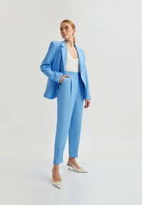 Lichtblauwe blazer en bijpassende op maat gemaakte broek met een gladde textuur. Witte top en crèmekleurige hakken met een driehoekig accent.