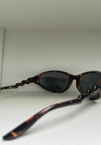 Lunettes de soleil en écaille de tortue avec verres foncés, présentant une monture lisse et courbée ainsi que des branches ondulées uniques. Protection CAT-3 indiquée.