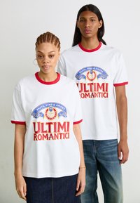Witte t-shirts met korte mouwen, voorzien van rode kraag en manchetten, met een kleurrijke gedrukte afbeelding die tekst en logo's bevat.