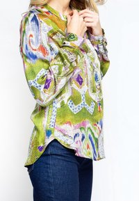 Kvinde iført en farverig bluse med abstrakt mønster og lange ærmer, i kombination med mørkeblå jeans, der retter kraven på blusen med hænderne.