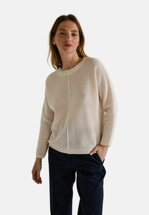 Femme portant un pull en tricot beige clair et un pantalon sombre, se tenant avec une main dans la poche devant un fond blanc uni.