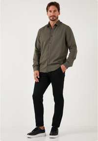 Camisa de manga larga verde oliva con cuello abotonado, combinada con pantalones ajustados negros y zapatillas negras. La camisa presenta una textura suave.