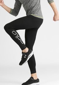 Leggings sportivi neri con una texture liscia, caratterizzati da un logo bianco prominente su un lato. Abbinati a scarpe sportive nere.