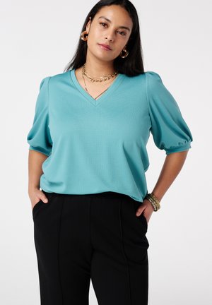 Vrouw met lang donker haar, gekleed in een teal V-hals top met pofmouwen, zwarte broek, gouden oorringen, gelaagde kettingen en armbanden, handen in de zakken.