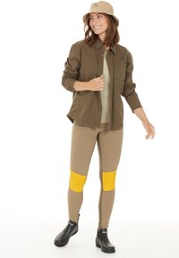 Brun knäppskjorta över en ljusgrön topp, tan leggings med gula knäfickor och svarta ankelskor; modellerad med en beige bucket hat.
