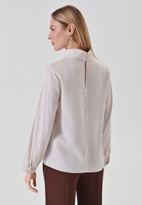 Helle beigefarbene Bluse mit hohem Kragen, langen Ärmeln und Knopfverschluss hinten. Glatte Textur mit leichtem Glanz, mit einem Schlitzdetail.