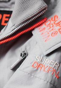 Detalje af lysegrå jakke med rød og hvid "Superdry JPN" broderi, en rød lynlås og en sort lynlåstrække med mærket "Superdry."
