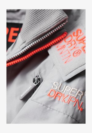 Veste grise avec une fermeture éclair orange, des logos brodés et un col texturé. Comprend une poche zippée et un tissu avec des lignes discrètes.