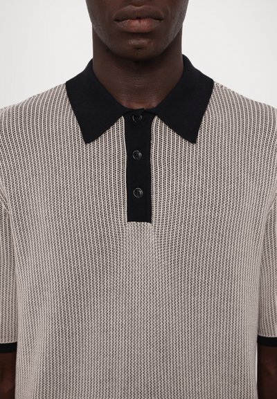 Polo en tricot beige avec un col bleu marine foncé. Présente un design texturé et côtelé ainsi que trois boutons noirs au niveau du col.