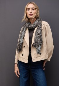 Beige dubbelzijdig colbert met zwarte knopen, gedragen over een donkerbruine top. Lange sjaal met luipaardprint hangt om de hals. Blauwe jeans.