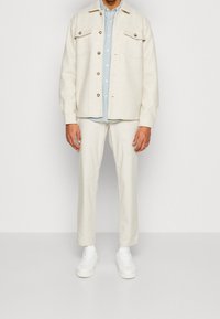 Giacca in misto lana chiara con tasche frontali, abbinata a una camicia button-up azzurro chiaro e pantaloni color crema, completata da sneakers bianche.
