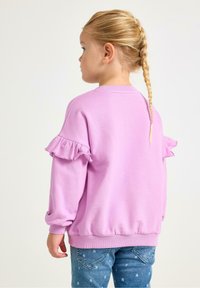 Felpa rosa chiaro con polsini a costine, caratterizzata da maniche arricciate e scollo rotondo. Abbinata a jeans blu decorati con motivi a forma di cuore.