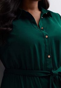 Emerald green button-up dress with a collar, featuring gold-tone buttons and a tied waist. Smooth fabric texture, no visible patterns.