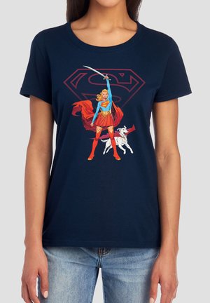 Woman wearing navy shirt featuring Supergirl with sword, red cape, and dog against large red Supergirl logo background.