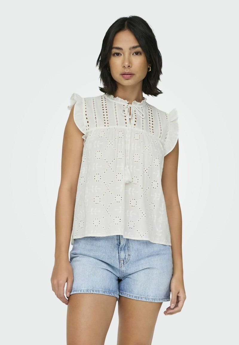 Blouse en coton blanc avec des manches volantées, des détails en broderie, un col à nouer et une coupe ample. Associée à un short en denim.