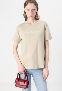 Beige bomull t-skjorte med korte ermer og rund hals, med hvit "Calvin Klein Jeans" tekst. Modellen holder en liten rød skinnevne.